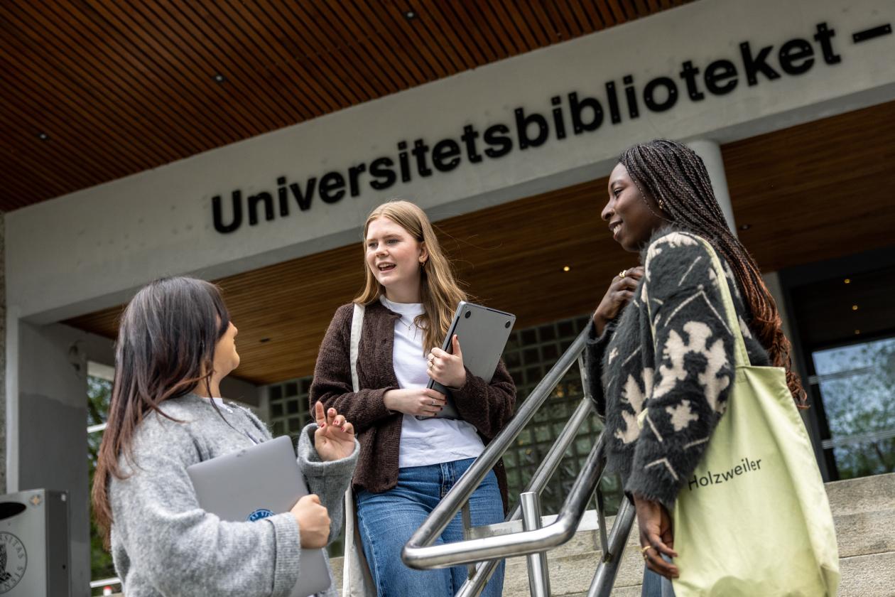 Tre unge kvinnelige studenter står i trappen til Universitetsbiblioteket og prater. 