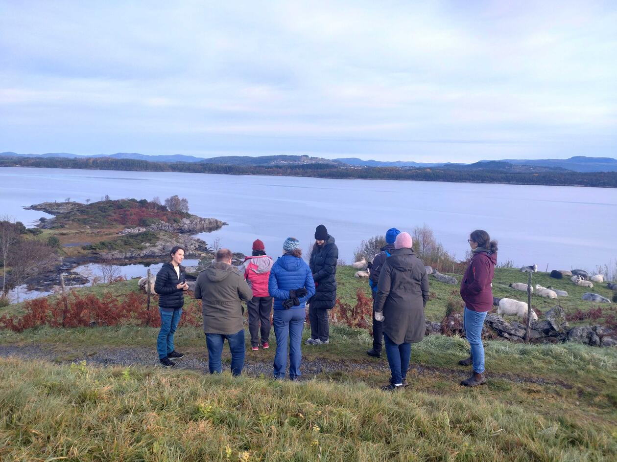 Group of people listening to woman talking, while enjoying the view of a fjord and heathlands