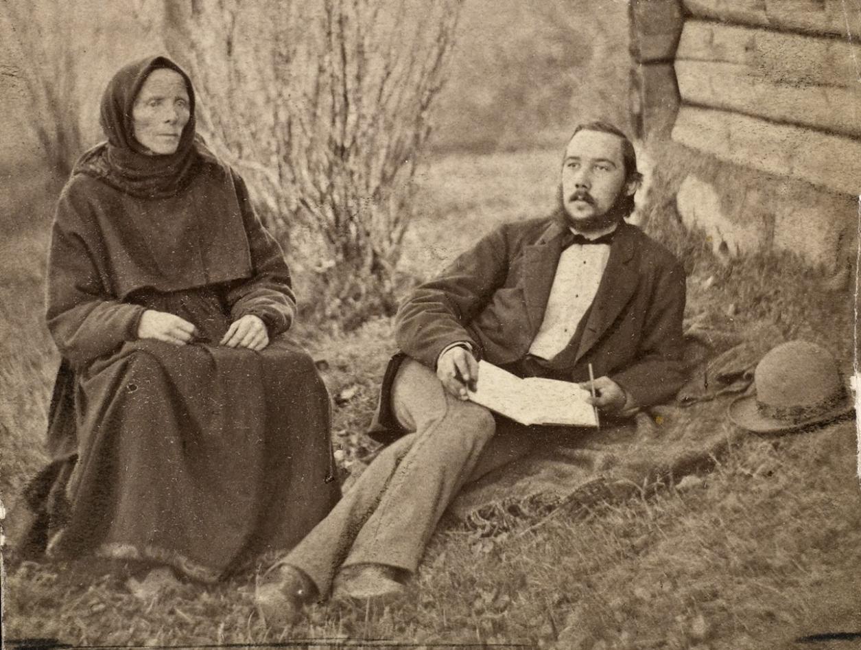 The picture is old in black and white. It shows a woman and a man outside. The woman sits with her hands in her lap staring blank forward. The man lay on the side with a book in his lap writing in the book, he stares out in the air.