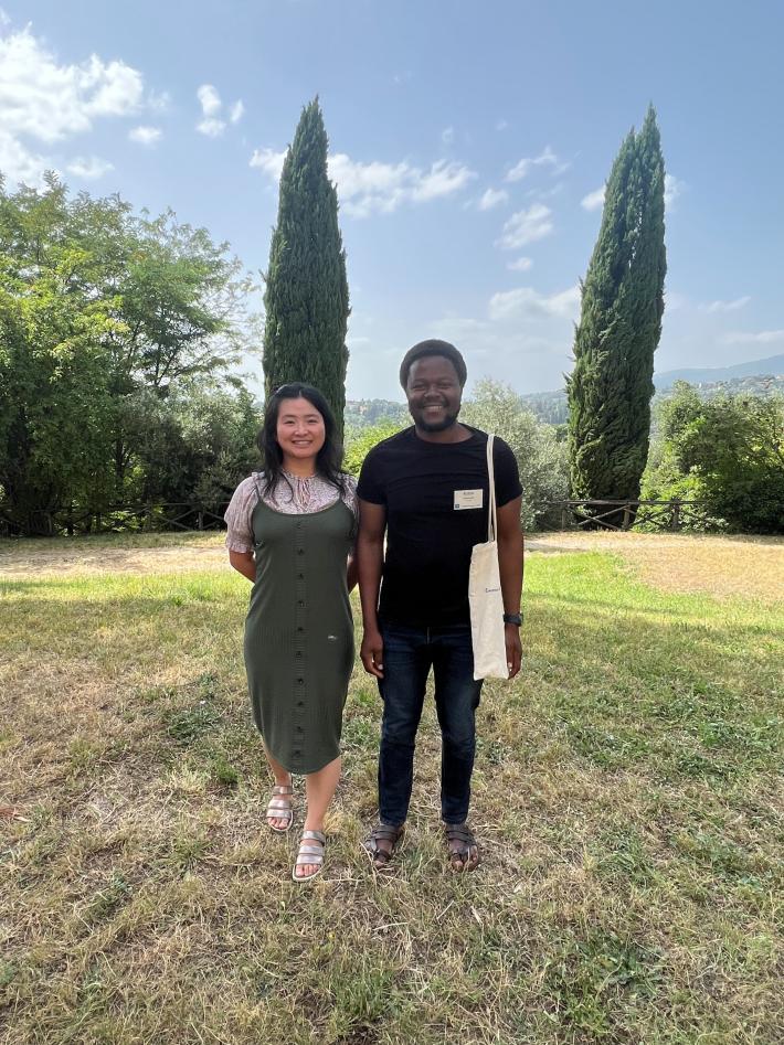 Two PhD students participating in a summer epidemiology course in Florence. On the left, a person smiles wearing a green dress; on the right, another smiles dressed in a black t-shirt and pants. They are posing in front of the campus garden.