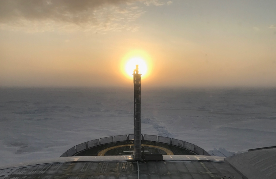 View from the research vessel Kronprins Haakon, overlooking a white sheet of ice with the sun just over the horizon in the center of the photo.