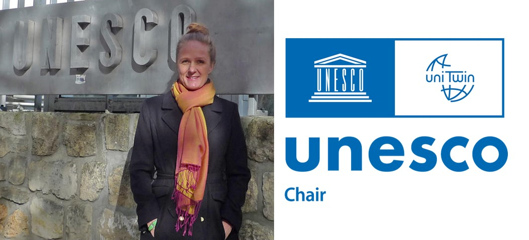 Smilende kvinne i kåpe og skjerf som står foran et stort UNESCO-skilt. Bildet er merket med UNESCO Chair-logoen