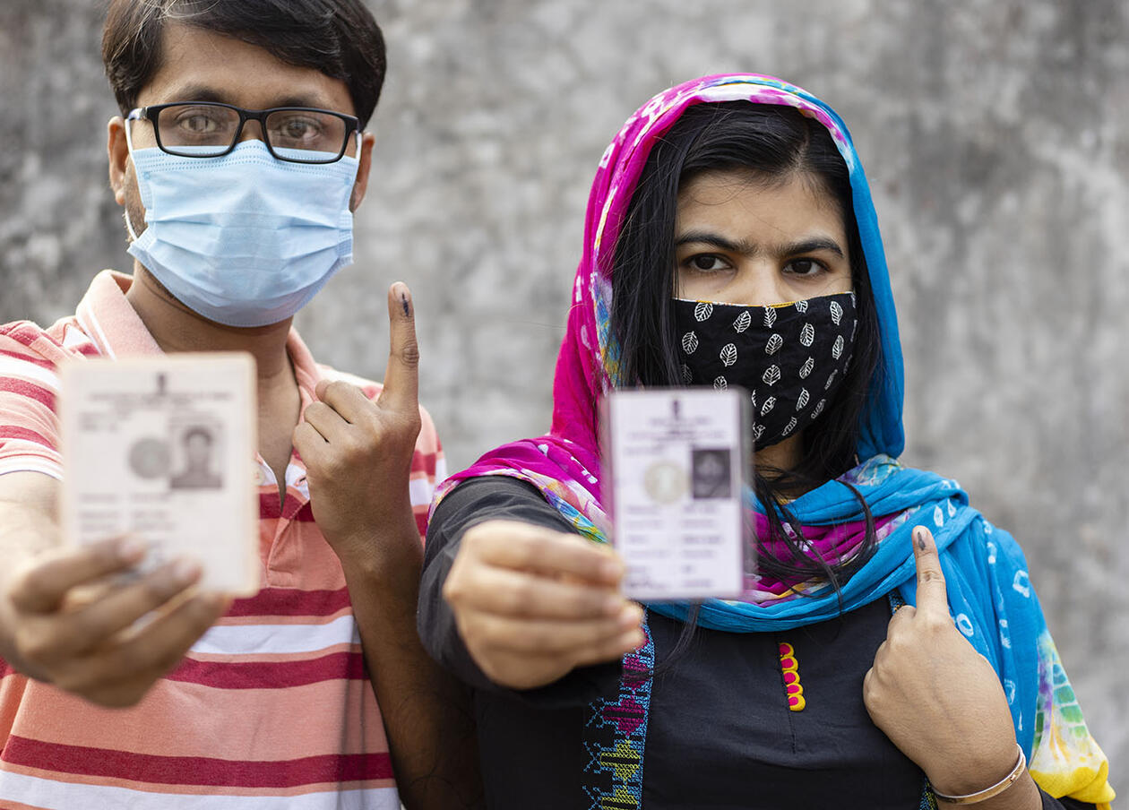 a man and woman showing ink-marked finger and voter card with safety nose mask on