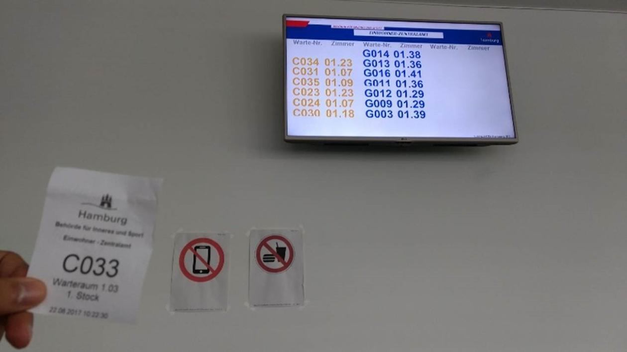 A screen with numbers on them in blue and yellow, and a hand holding up a queue ticket
