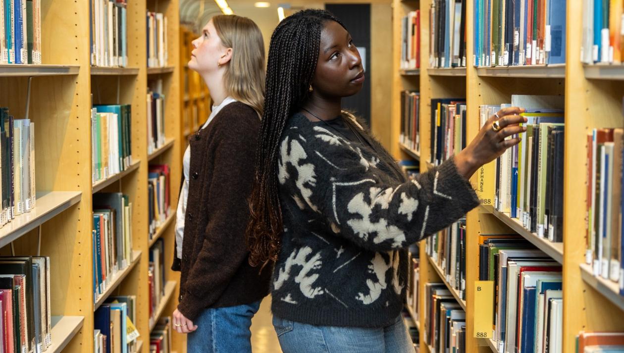 To jenter/studenter står rygg i rygg mellom to hyller på biblioteket og leiter etter bøker. 