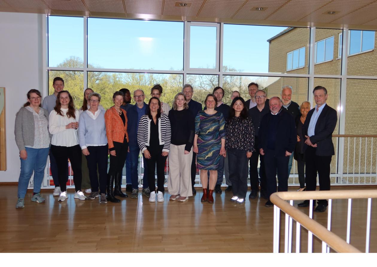 Researchers from the Respiratory Health in Northern Europe (RHINE) study consortium at Aarhus University on April 16, 2024. They are standing inside a campus building, in front of a large window with a view of green trees and a blue sky.