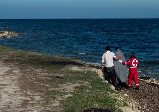 Tre mennesker går langs en strand, en person har et grått teppe over seg og er flankert i en person i Røde Kors-uniform på høyre side og en mann i hvit skjorte på venstre side.