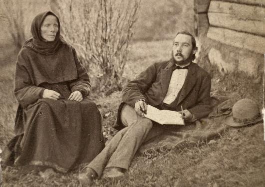 The picture is old in black and white. It shows a woman and a man outside. The woman sits with her hands in her lap staring blank forward. The man lay on the side with a book in his lap writing in the book, he stares out in the air.