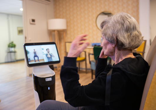 An old lady sitting in a chair, following a video on a screen on a robot on how to exercise while sitting.