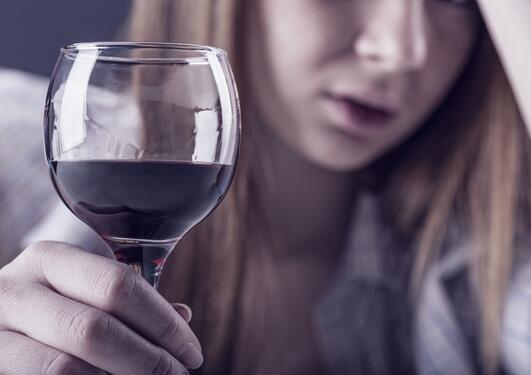Kvinne sitter med hånden i hodet, har et glass rødvin foran seg. Ser oppgitt ut sliten ut