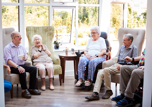 Four elderly people in a nursing home talking, laughing and drinking cooffee, all have smartwatches.