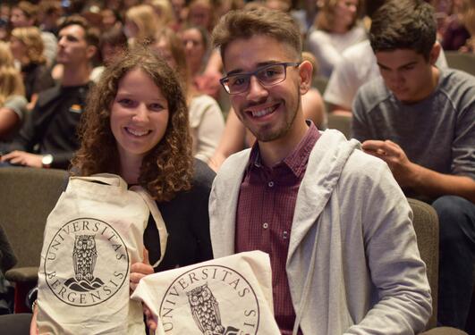 Two students smiling, facing the camera, holding a UiB tote bag