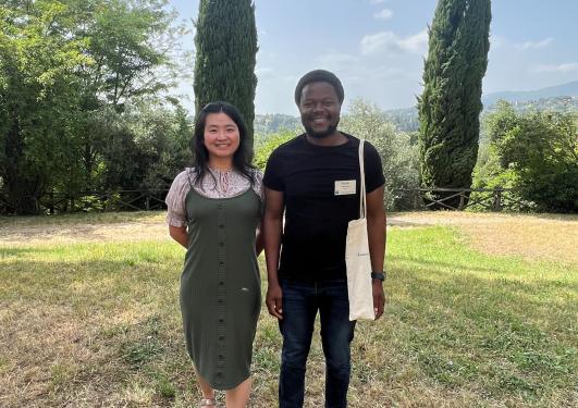 Two PhD students participating in a summer epidemiology course in Florence. On the left, a person smiles wearing a green dress; on the right, another smiles dressed in a black t-shirt and pants. They are posing in front of the campus garden.