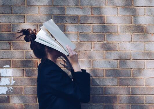 Student in front of natural rustic red brick background holding book up to her face.