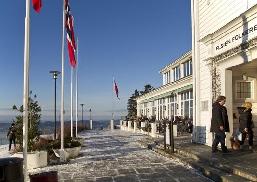 A white building to the right, snow-clad ground, blue sky and several Norwegian flags on poles, sea and mountains in the background