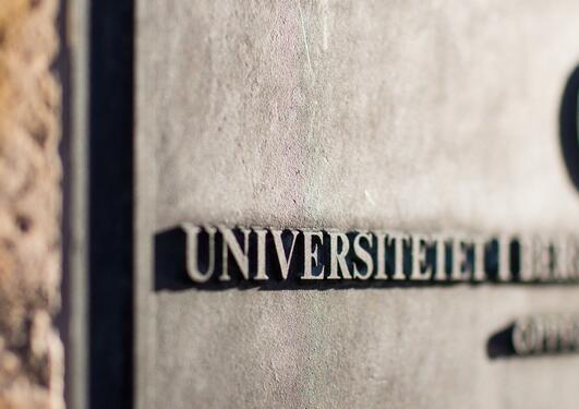 Bilde av en plakett på en murvegg med logoen og navnet til Universitetet i Bergen hvor halve bildet er ute av fokus
