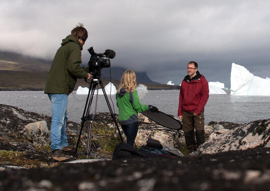 To personer filmer mann som står foran havet med isbreer i bakgrunnen