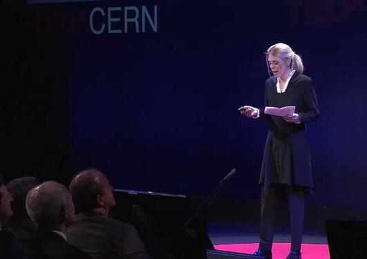 A blonde woman in black on a stage, some people at the front, the word CERN can be seen behind her, in white on a dark background