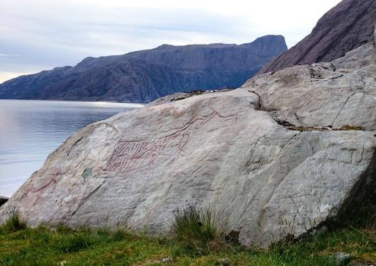 Berg med rødmalte helleristninger på. Fjord og fjell i bakgrunnen.