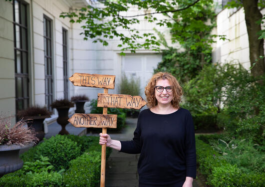 Zhanna Gaulen står i musehagen med et skilt hvor det står "this way" og "that way" som peker i ulike retninger. Frodig grønn bakgrunn, hun har sort genser, rødt krøllete hår og briller og smiler inn i kameraet 